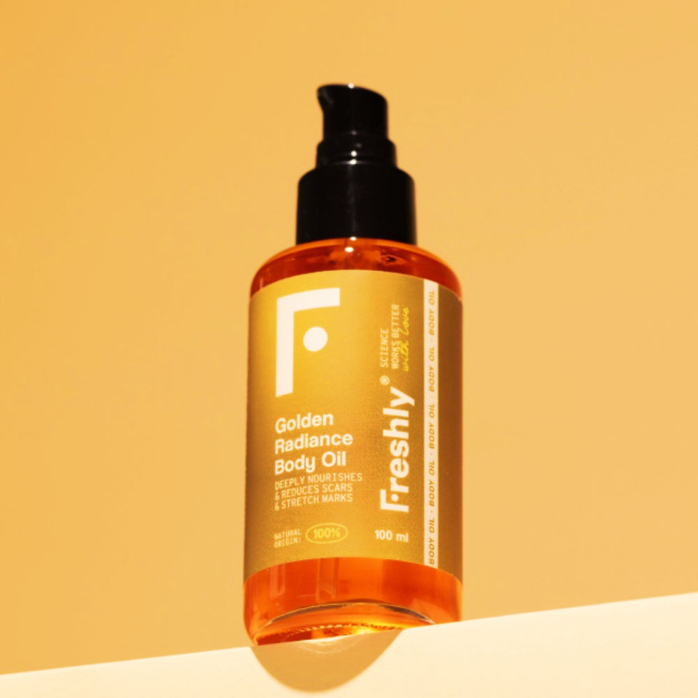 Frasco de 100ml de Freshly Óleo Corporal de Brilho Dourado com bomba em pé sobre fundo amarelo. O frasco âmbar possui rótulo amarelo e branco, oferecendo nutrição corporal profunda para cuidado corporal natural.